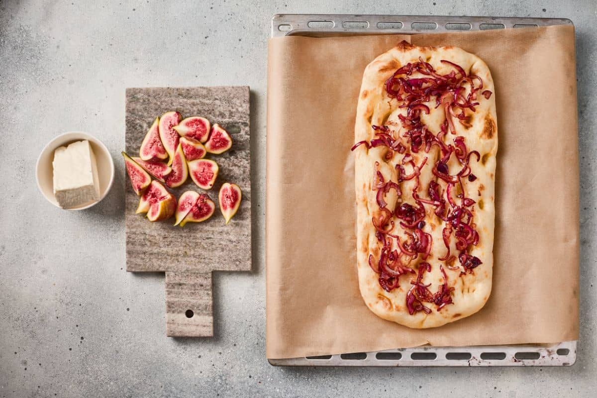 Flatbread on a tray lined with parchment paper, with a fig slice placed on a cutting board and other ingredients on the side.