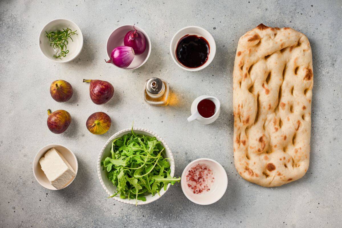Flatbread ingredients arranged on a surface, including figs, caramelized onions, goat cheese, herbs, and olive oil.