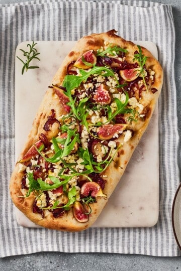 Served flatbread garnished with arugula and goat cheese, surrounded with ingredients on a tablecloth underneath.