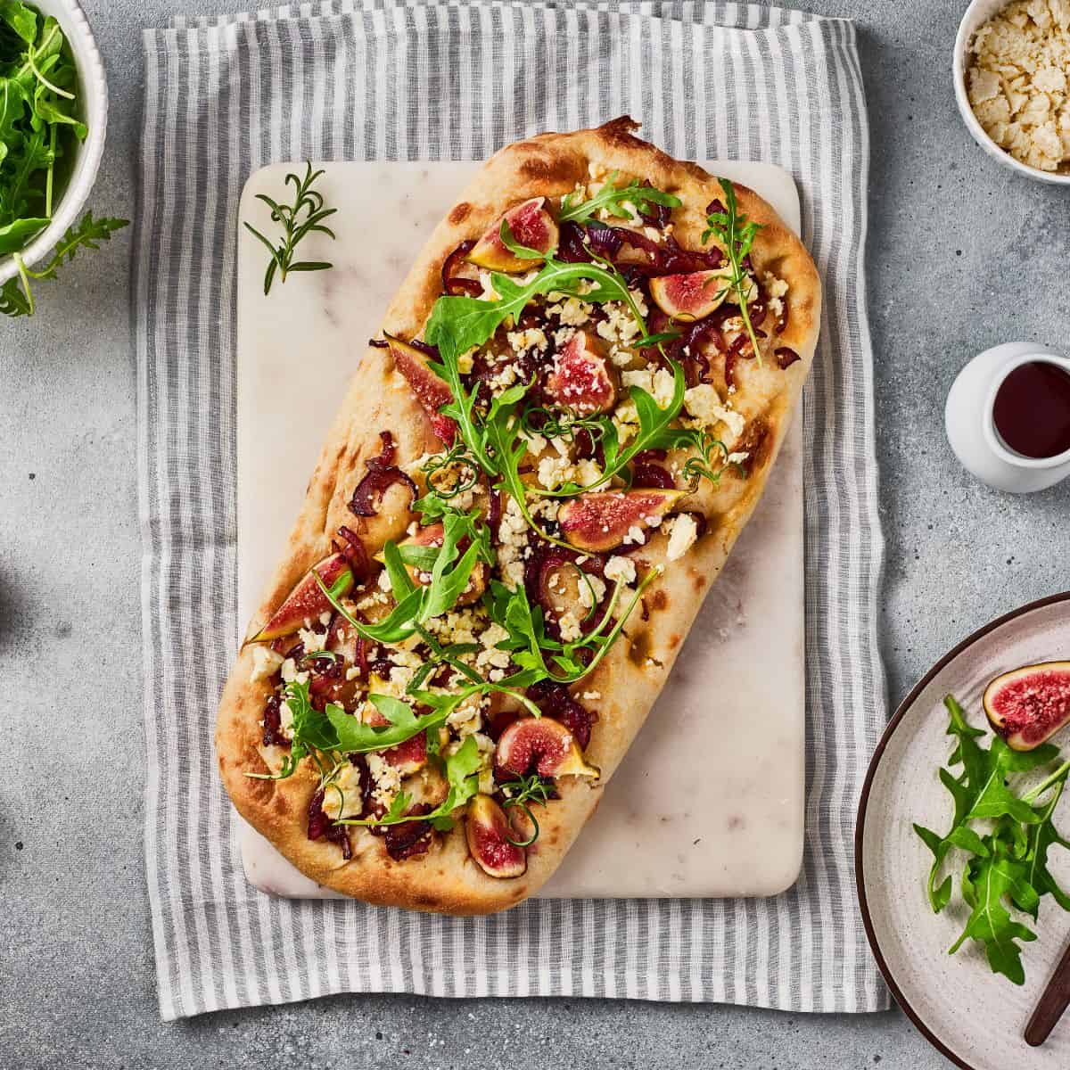 Served flatbread garnished with arugula and goat cheese, surrounded with ingredients on a tablecloth underneath.