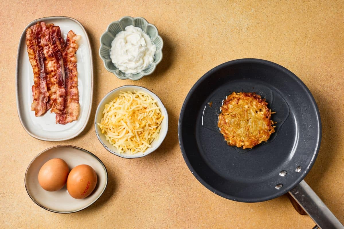 Fully loaded hashbrowns placed in a pan, surrounded by ingredients like grated potatoes, cheddar cheese, bacon, eggs, green onions, and sour cream.