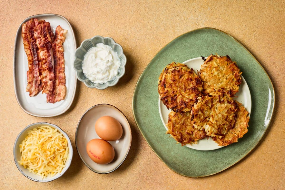 Fully loaded potato hashbrowns on a plate beside the ingredients, cheddar cheese, bacon, eggs, green onions, and sour cream.
