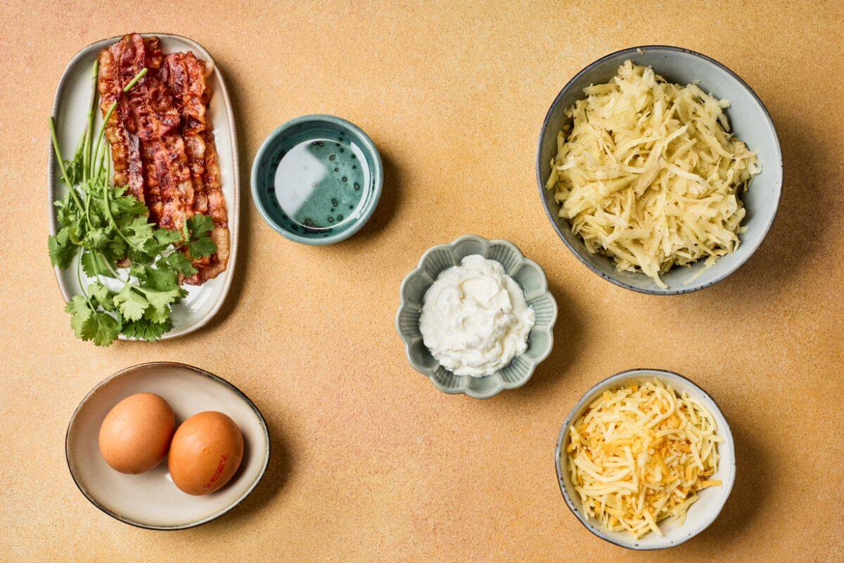Ingredients for fully loaded hashbrowns arranged on a surface, including grated potatoes, shredded cheddar, bacon, eggs, green onions, and sour cream.