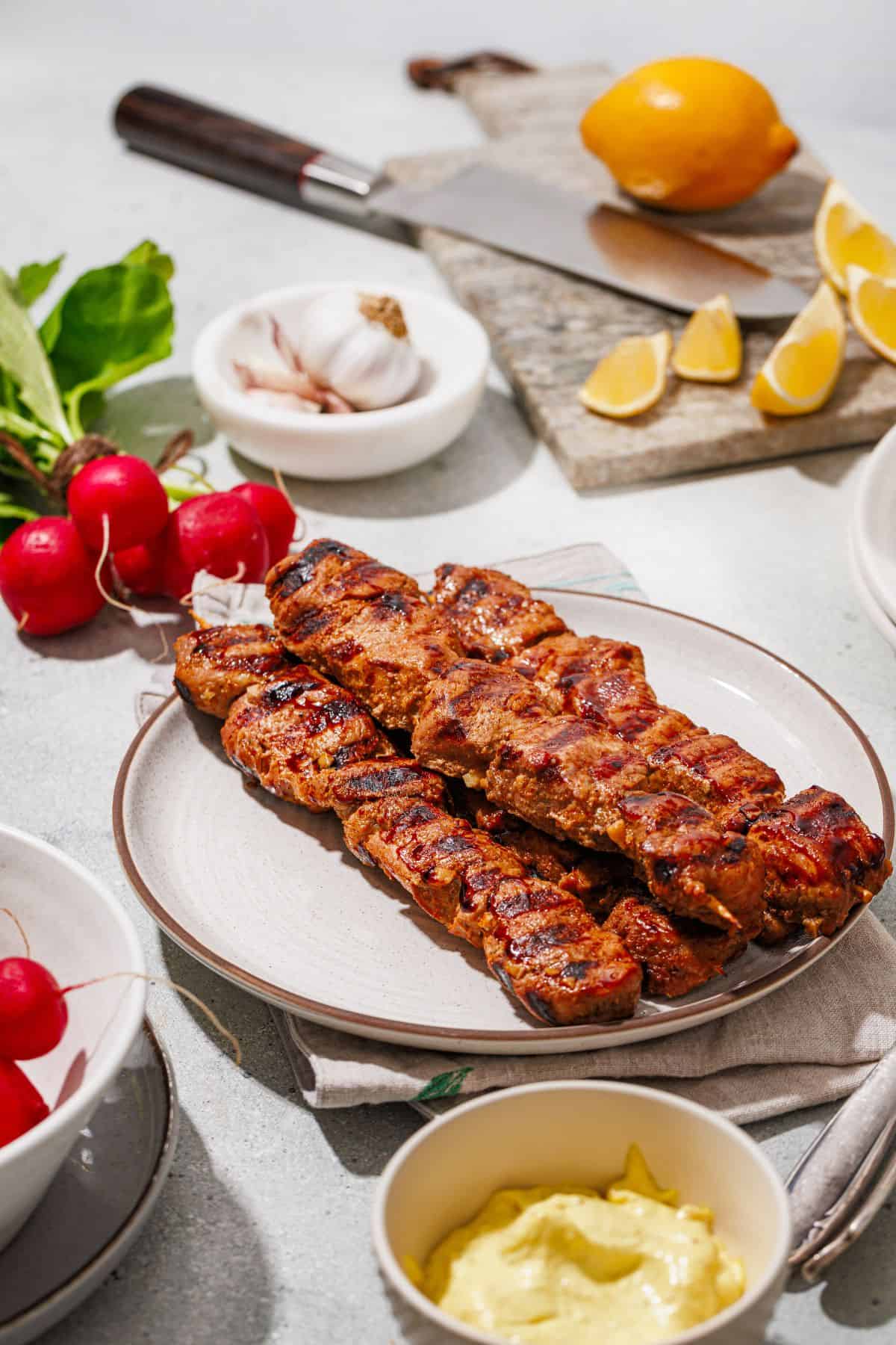 Grilled marinated pork skewers with homemade aioli served on a plate, surrounded by cherry tomatoes and sliced lemons.