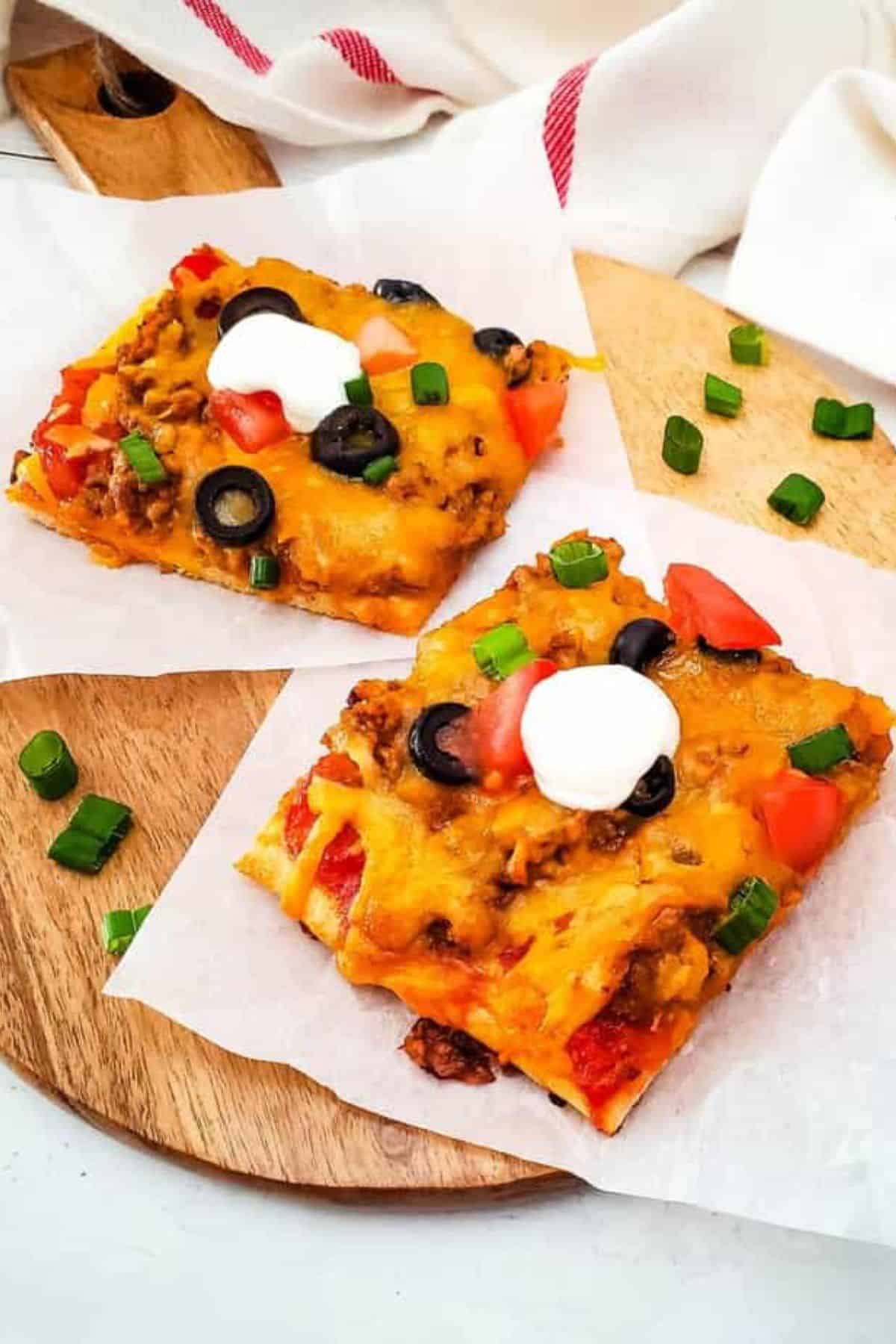 Two slices of taco pizza on parchment paper, served in a wooden serving board.