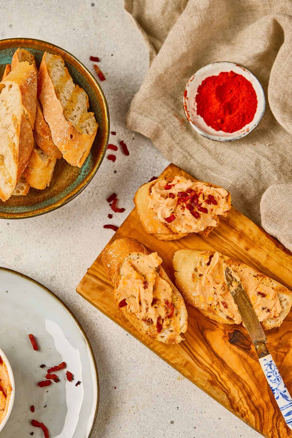 A small bowl of maple bacon cheese dip on a wooden board, with garlic bread beside it and a knife spreading the dip.