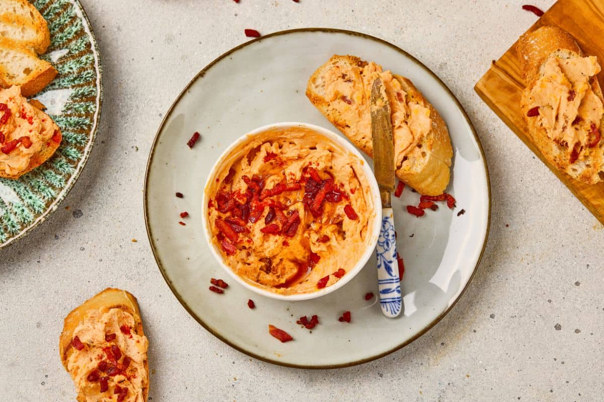 A small bowl of maple bacon cheese dip placed on a plate next to garlic bread, with a knife spreading the dip.