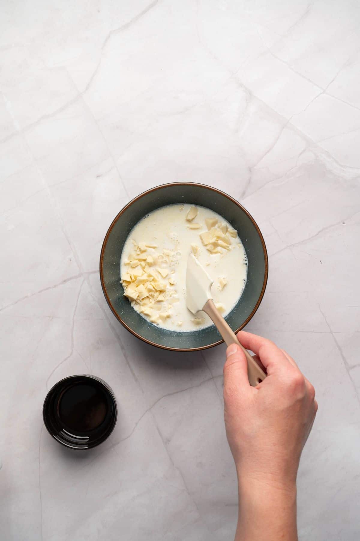 A hand holding a spatula mixing a small bowl of pistachio cheesecake filling.