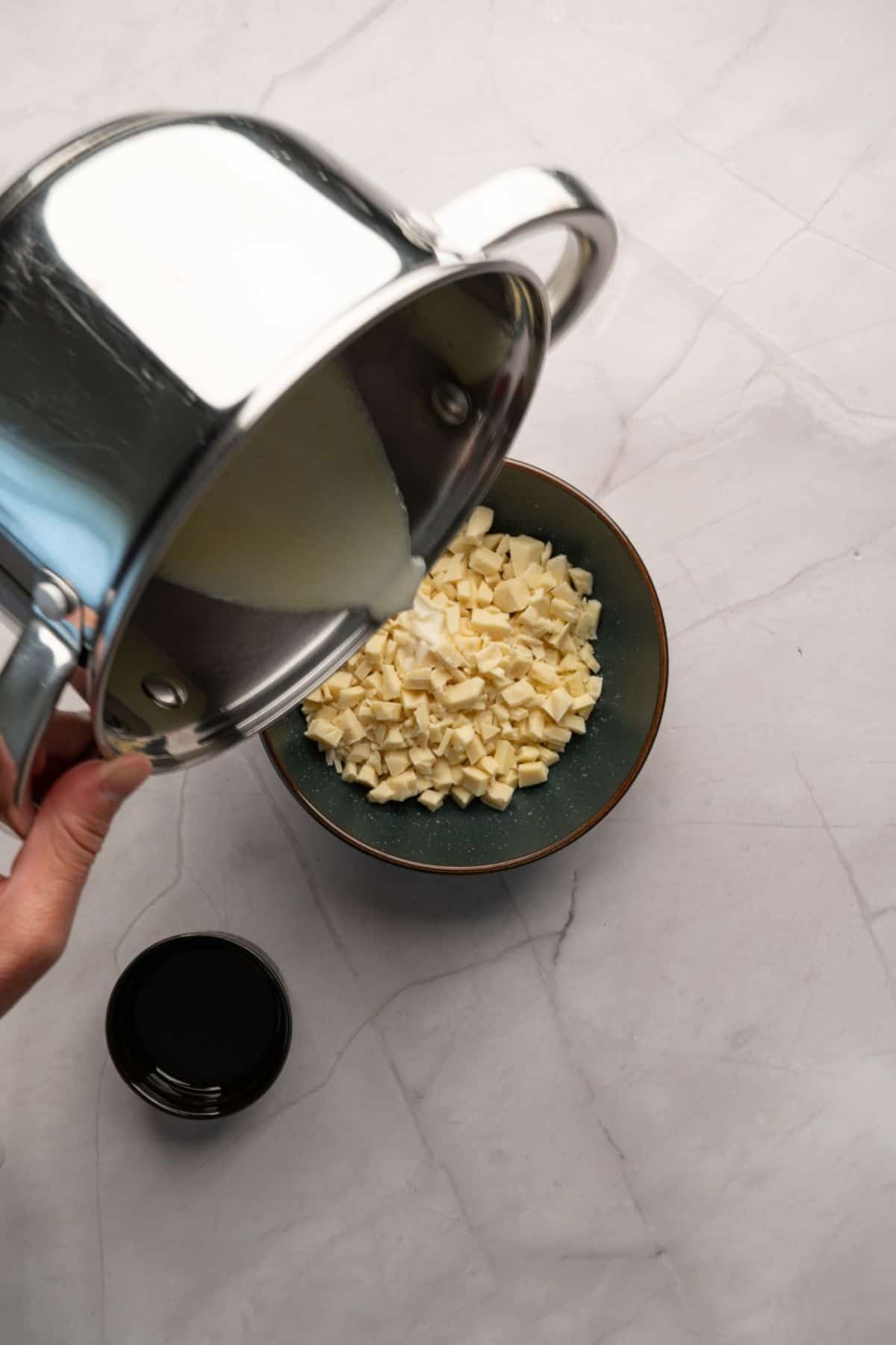 A hand pouring a pot of warm cream into a bowl of chopped white chocolate.