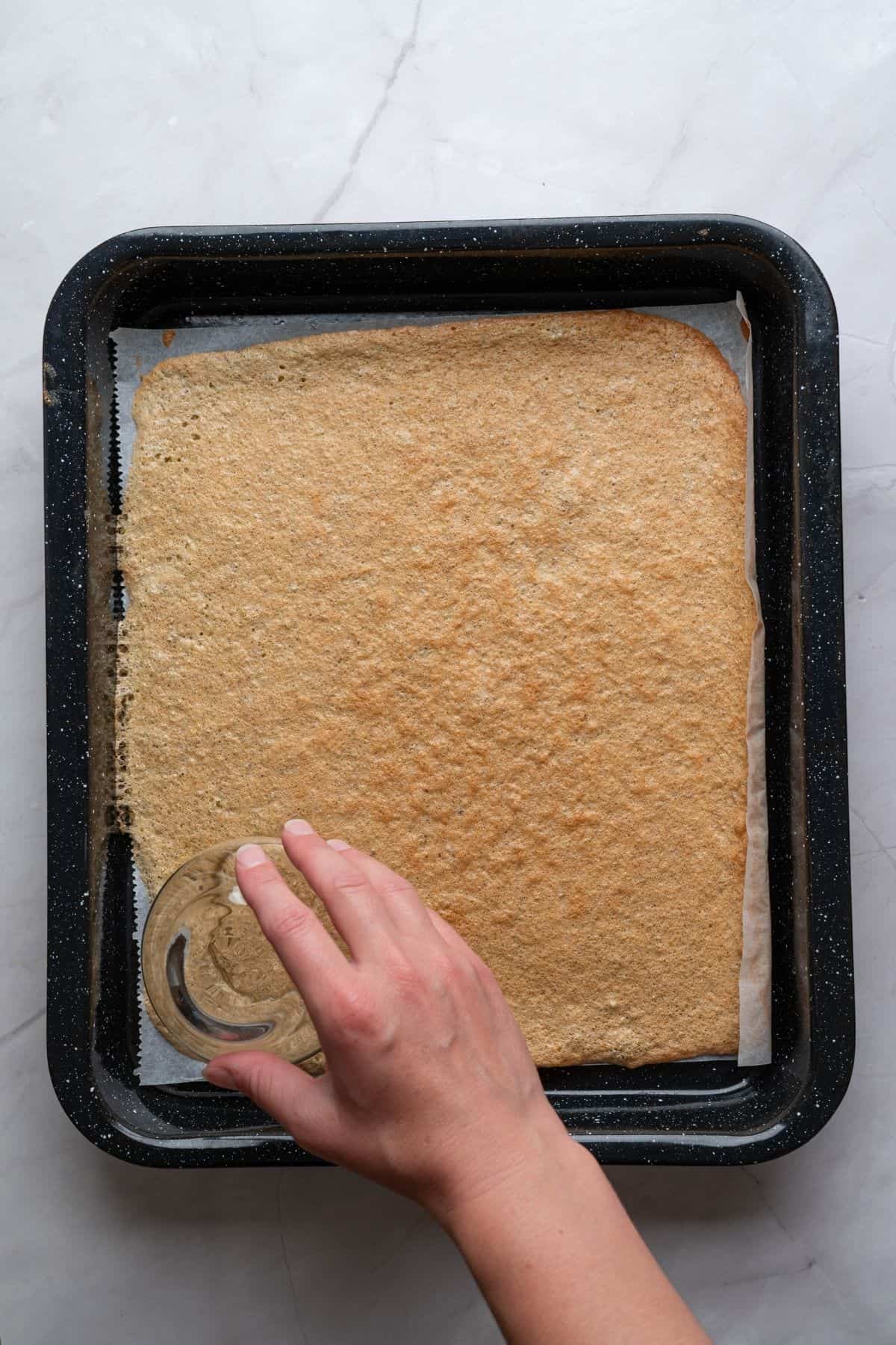 A hand holding a glass, shaping the pistachio sponge layer on a baking sheet.