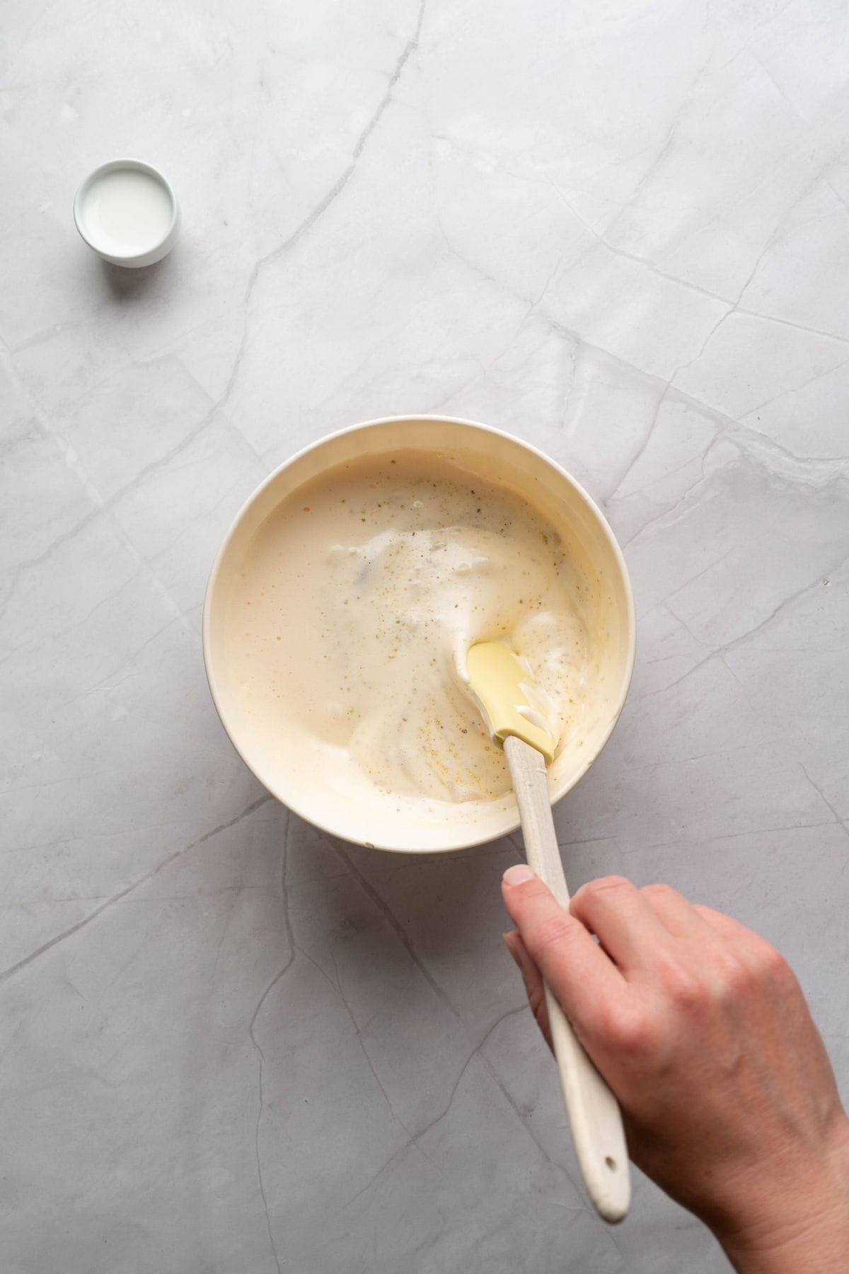 A hand holding a spatula, mixing pistachio cheesecake filling in a bowl.