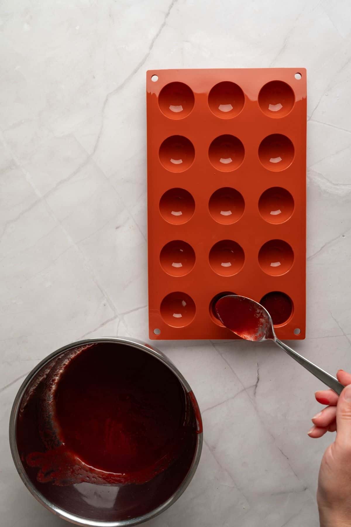 A hand pouring raspberry gelée mixture into small dome molds.