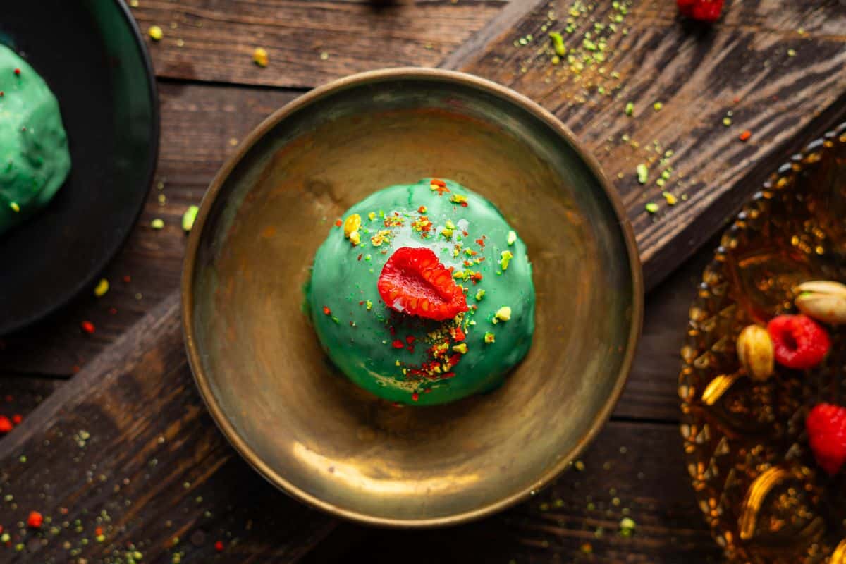 A dome-shaped pistachio cheesecake on a plate, surrounded with fresh raspberries for garnish.