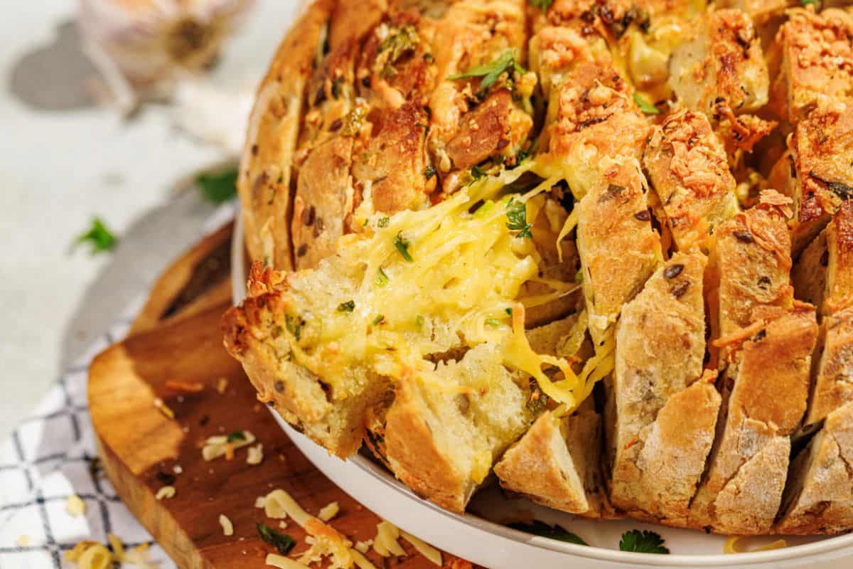 A golden, cheesy pull-apart garlic bread, partially sliced and garnished with fresh parsley, served on a plate placed on a wooden board, with melted cheese oozing between the cuts.