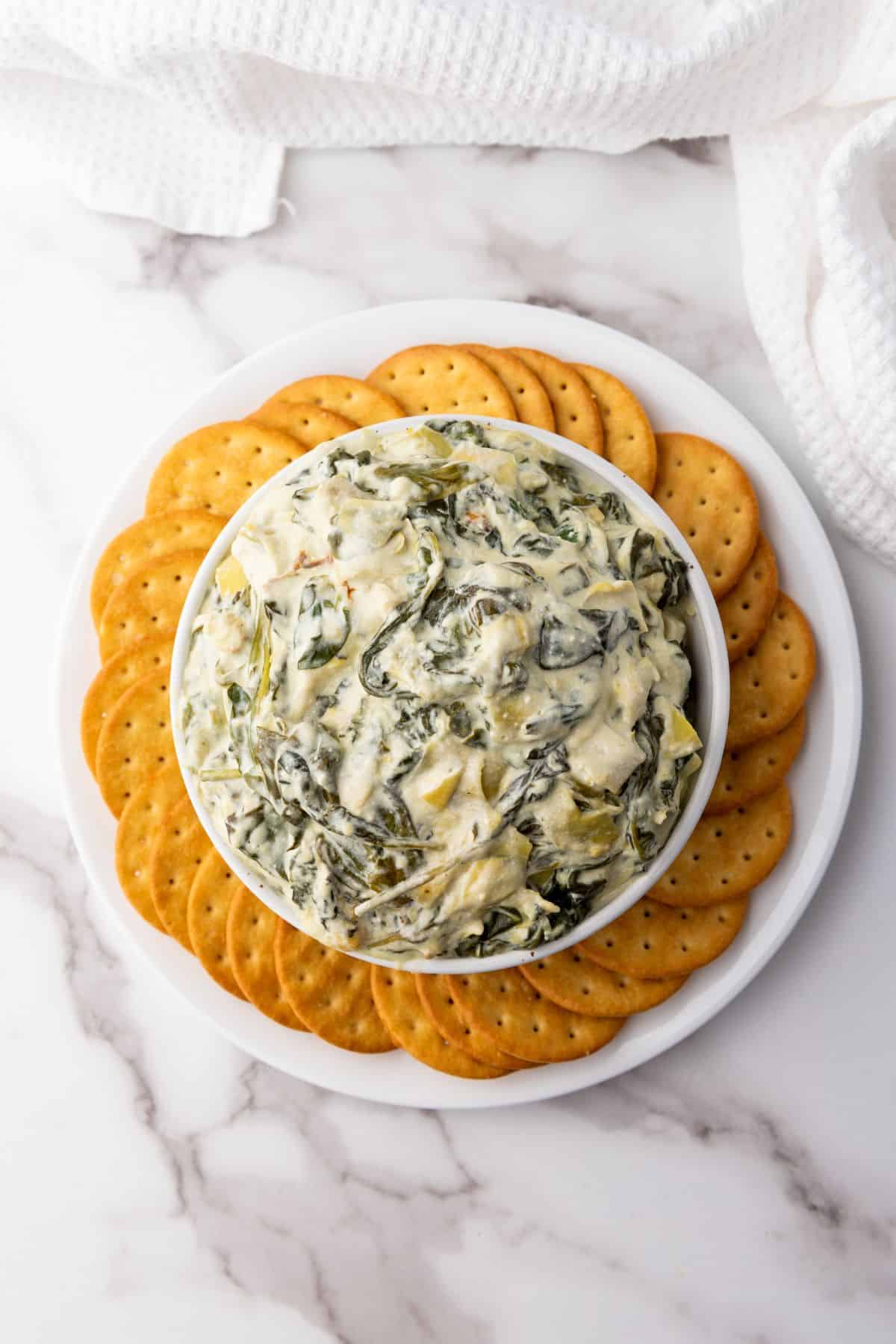 Creamy artichoke dip served in a bowl and surrounded by crisp biscuit crackers for dipping.