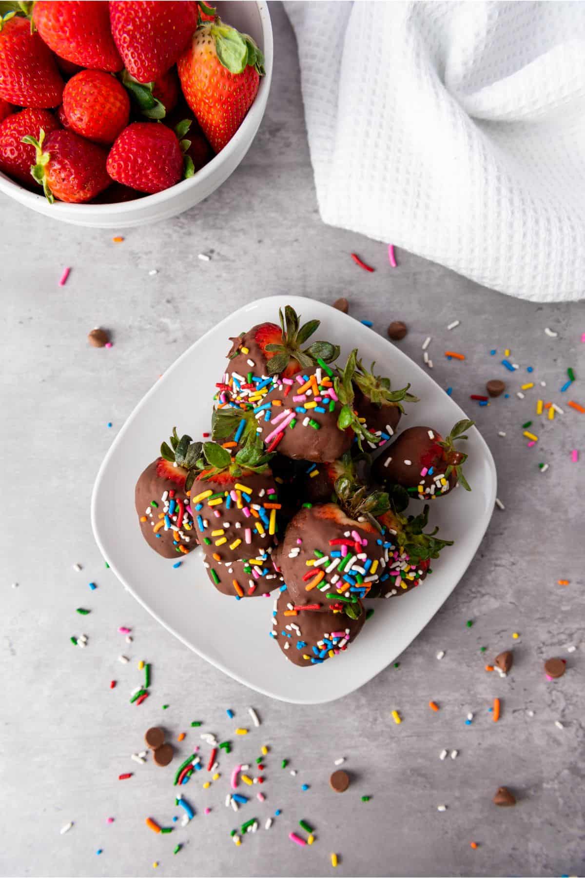 Close-up of chocolate-dipped strawberries with sprinkles on a white dish. Fresh strawberries in a bowl and colorful confetti scattered on the table.