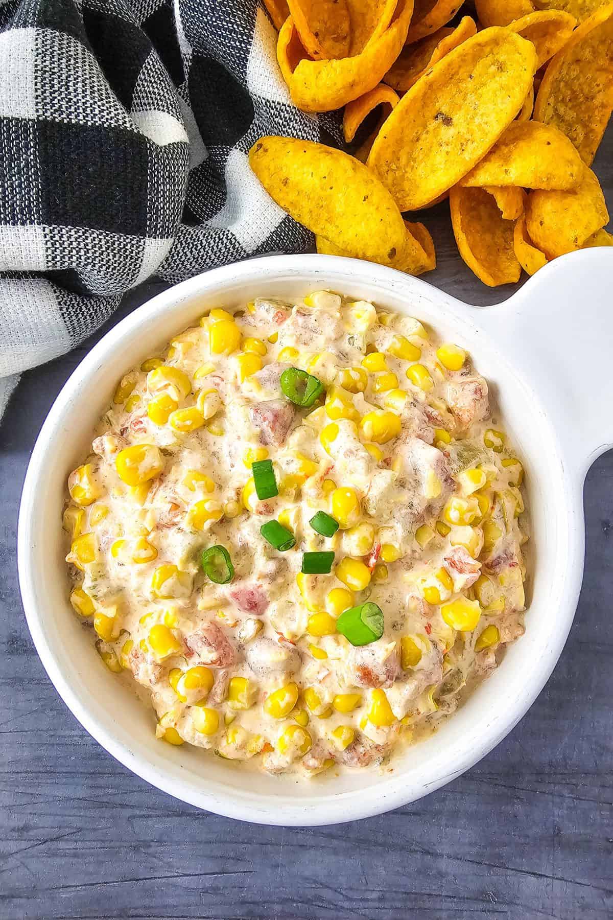 Creamy corn salad with diced vegetables served with crispy tortilla chips on a dark wooden surface.