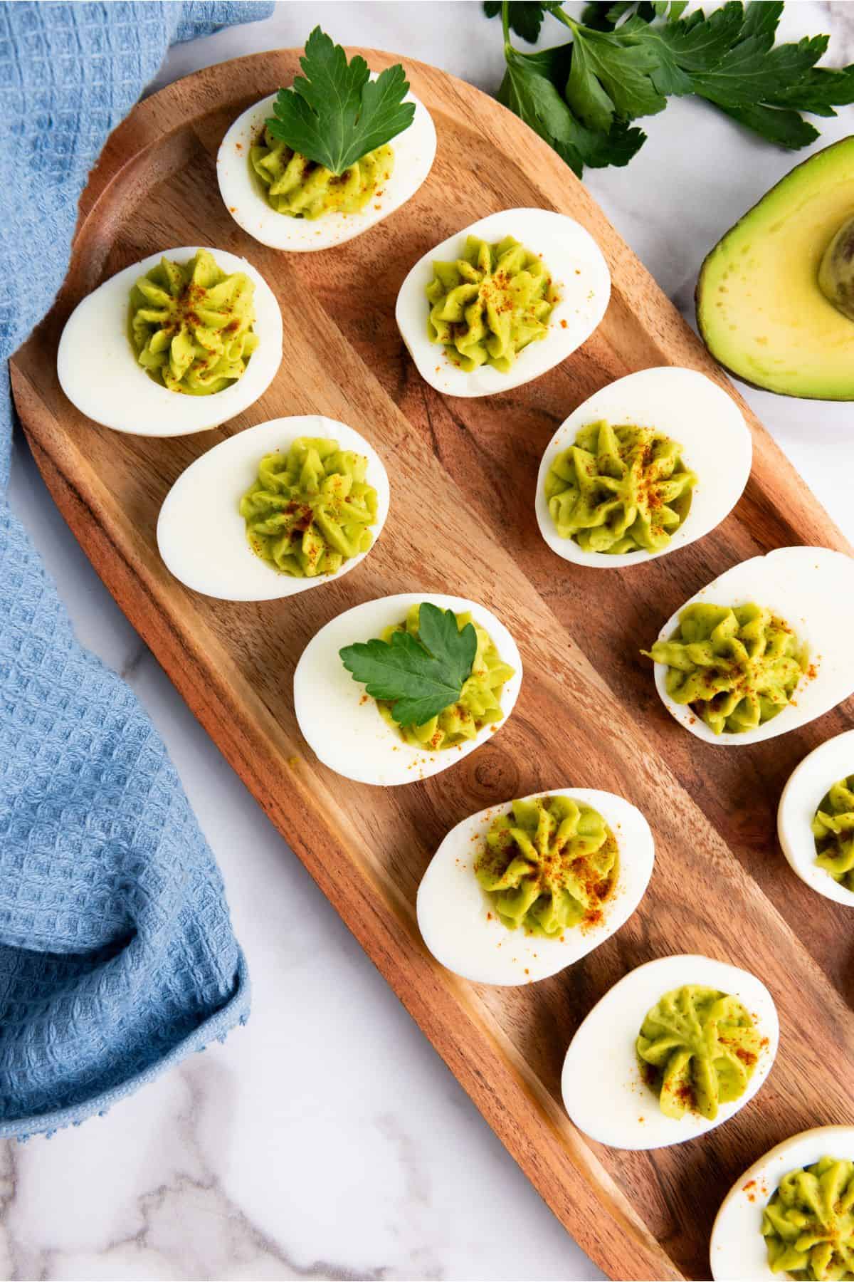 Creamy deviled eggs topped with avocado, garnished with parsley on a wooden tray.