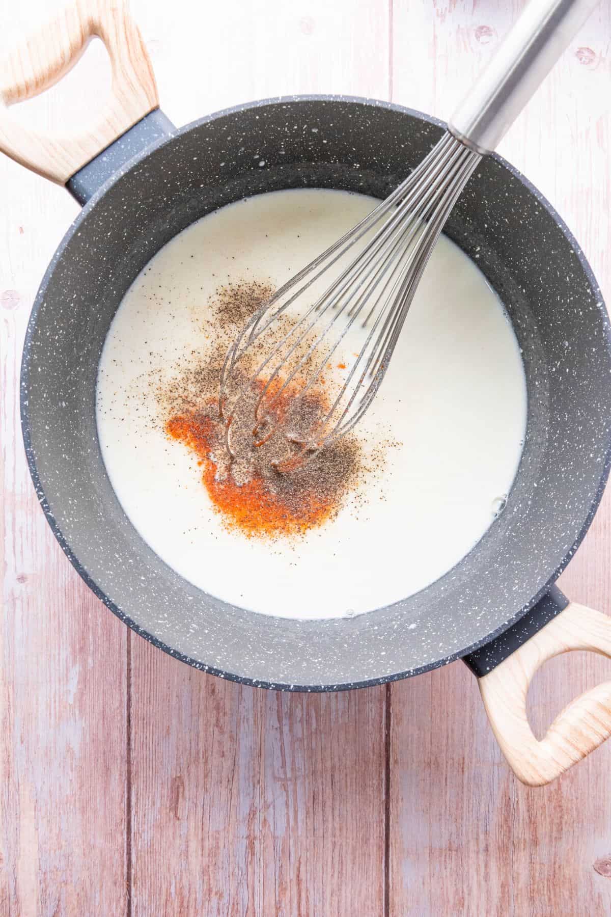 Creamy broth with spices in a black saucepan on a pink wooden surface, whisk for cooking.