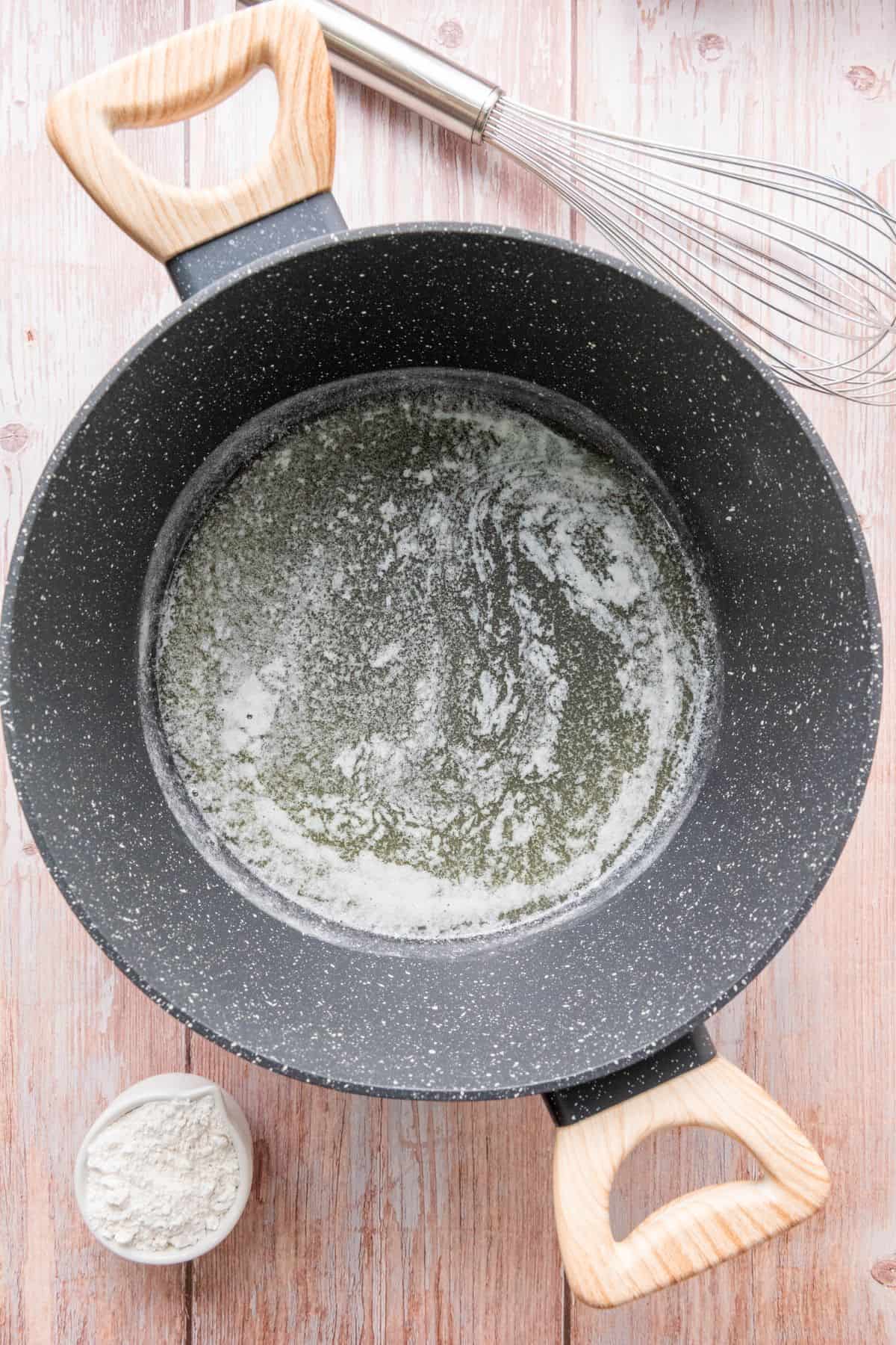 A black cooking pan with melted butter, a whisk, and a small bowl of flour on a wooden countertop.