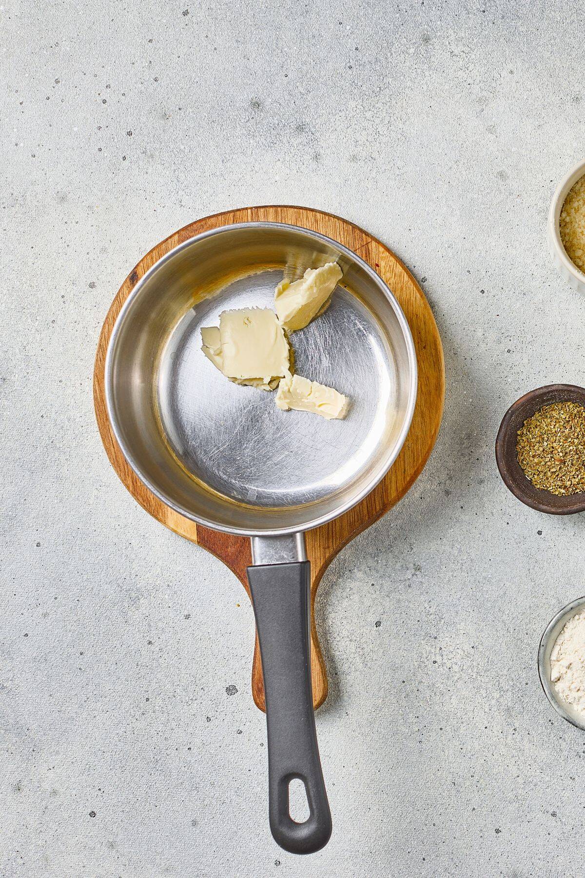 Small saucepan with melting butter on a wooden board, surrounded by spice bowls, on a gray countertop.
