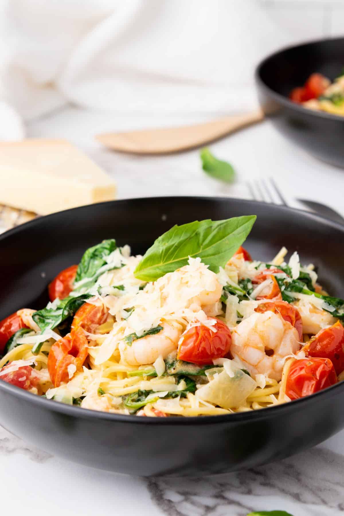 Delicious seafood pasta with cherry tomatoes, shrimp, basil leaves, and cheese in a black bowl.