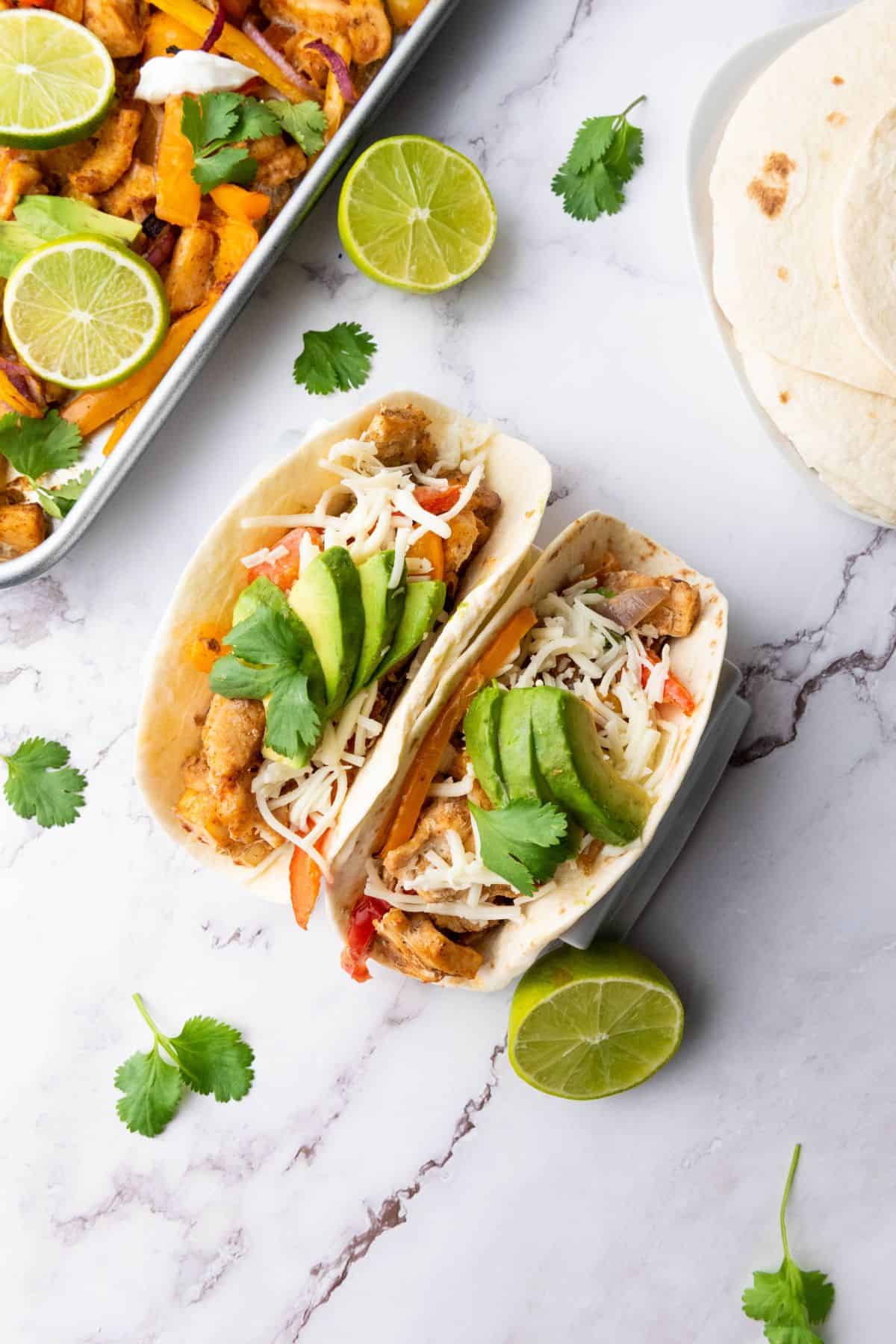Vibrant tacos filled with chicken, cheese, avocado, and fresh herbs on a white marble surface.