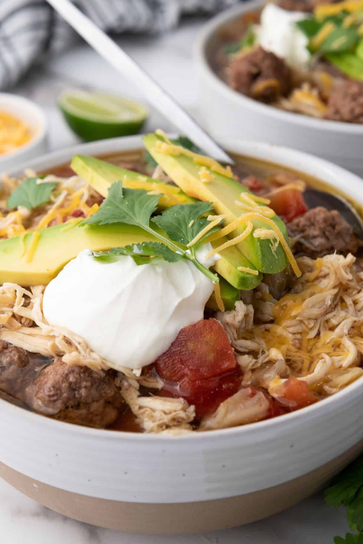 Close-up of a hearty chicken and vegetable soup with avocado, shredded cheese, sour cream, and fresh cilantro on top.