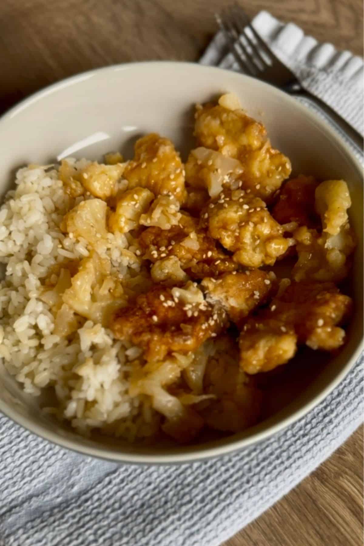 A bowl of chicken with sesame seeds served over rice, garnished with sauce, on a textured cloth with forks.