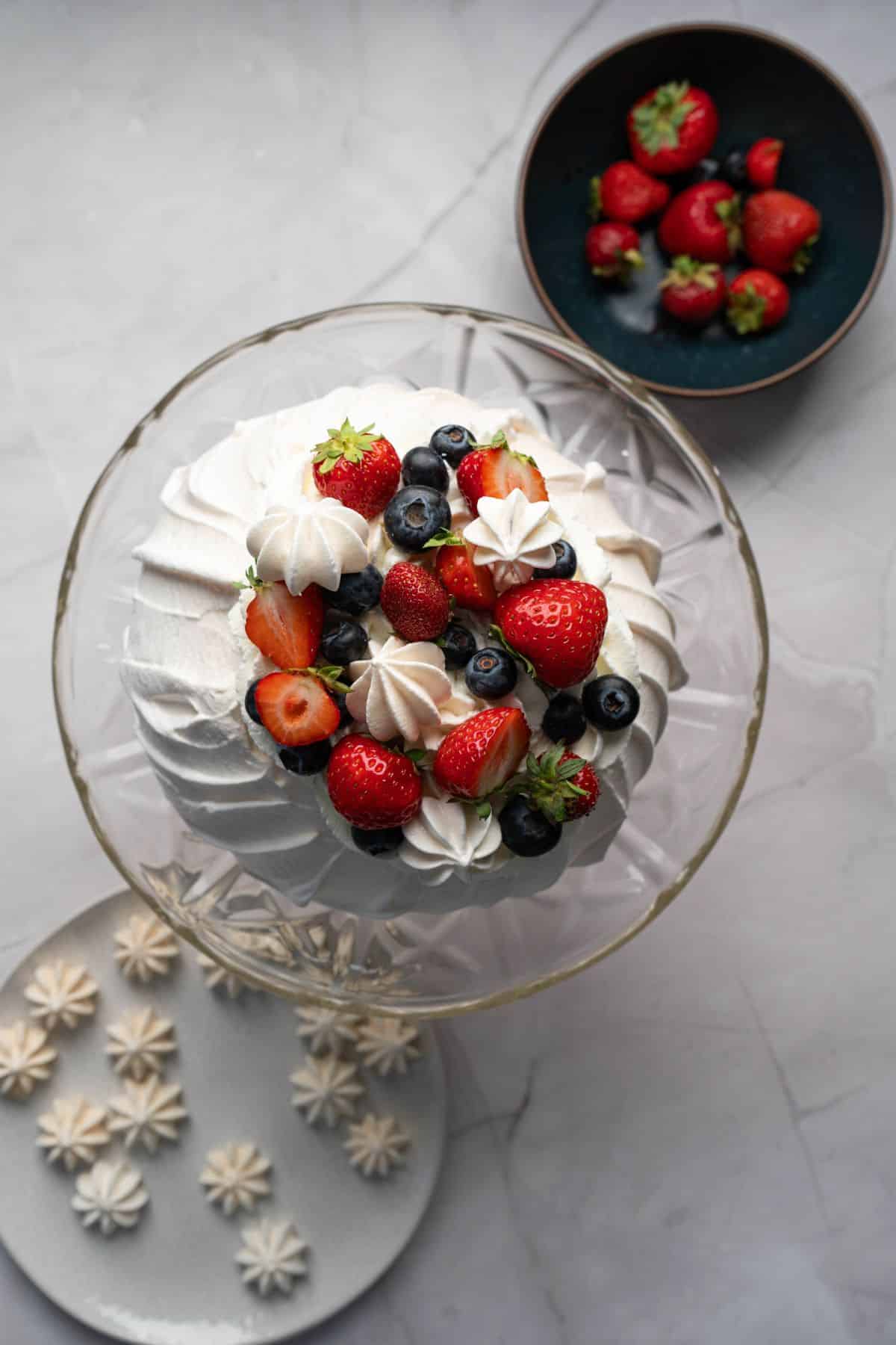 Fresh berries on a cake with whipped cream and meringue, with additional berries in a bowl nearby.
