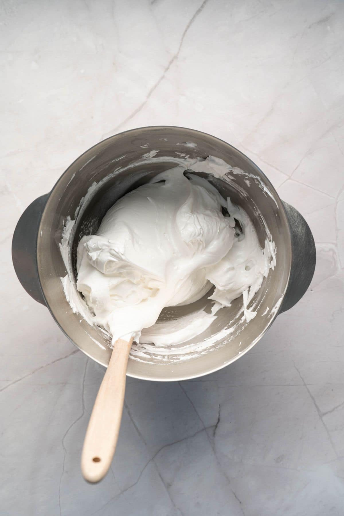 Silky white whipped cream in a stainless steel bowl with a wooden spatula.