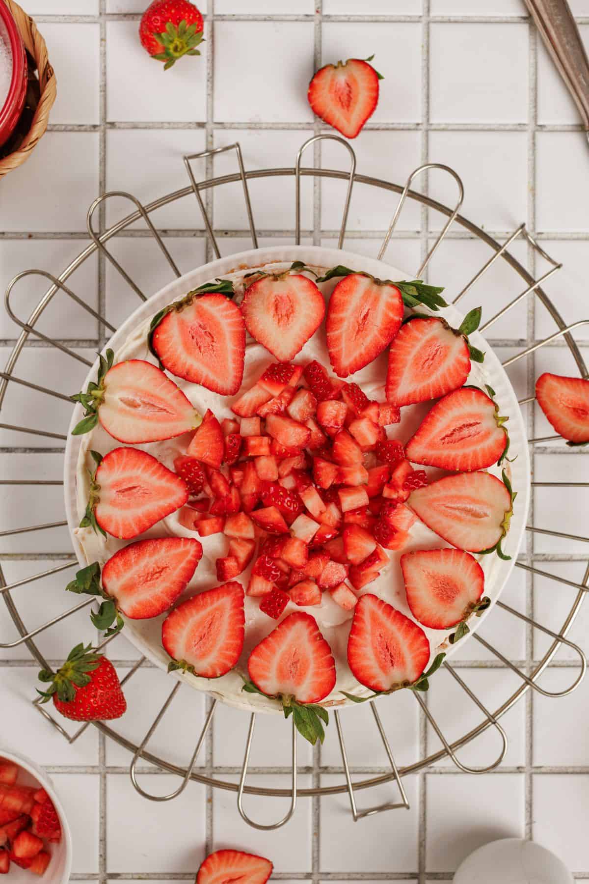Close-up of a strawberry cake topped with fresh sliced strawberries, served on a wire rack on a white tiled surface.