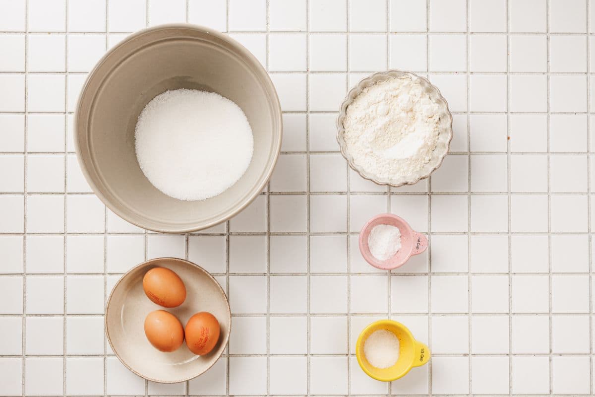 A variety of baking ingredients including eggs, flour, sugar, and baking powder on a white tiled surface.