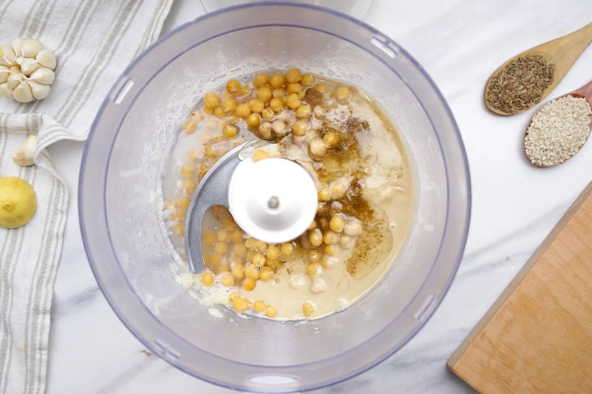 A food processor blending chickpeas, tahini, garlic, and spices to make smooth hummus, with ingredients and utensils visible.