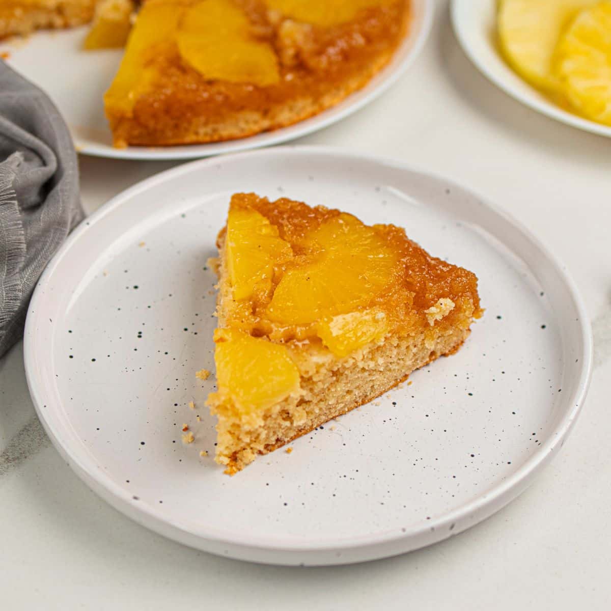Delicious pineapple upside-down cake slice on a white speckled plate, perfect for dessert or afternoon treat.