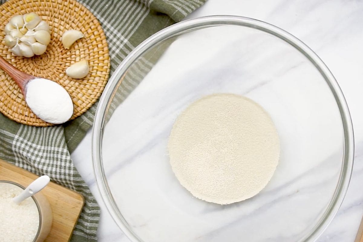 Fresh garlic bulbs and peeled cloves on a wooden board and a woven tray, with a bowl of grated garlic on a marble surface.