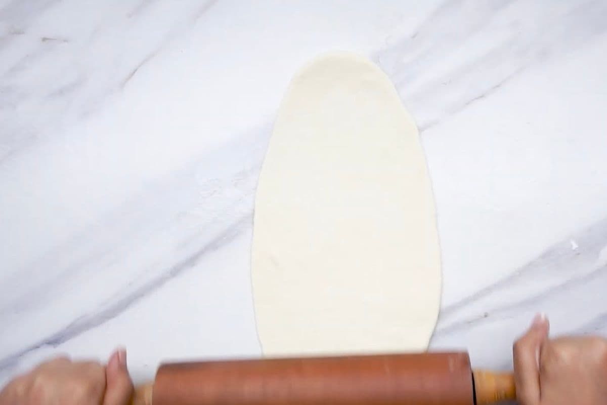 A person uses a wooden rolling pin to evenly flatten dough on a marble countertop.