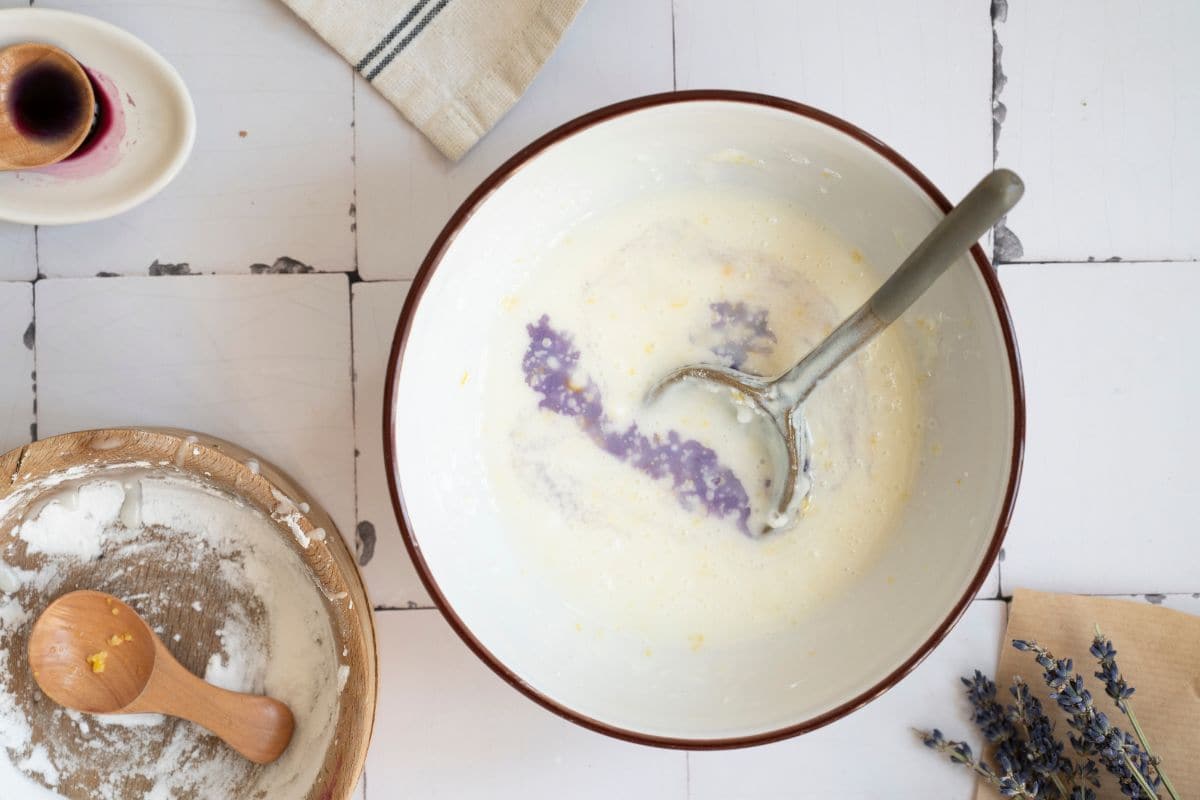 A bowl of creamy cake batter with vanilla and purple food coloring, ready for baking. Whisk and ingredients visible, rustic white table setup.