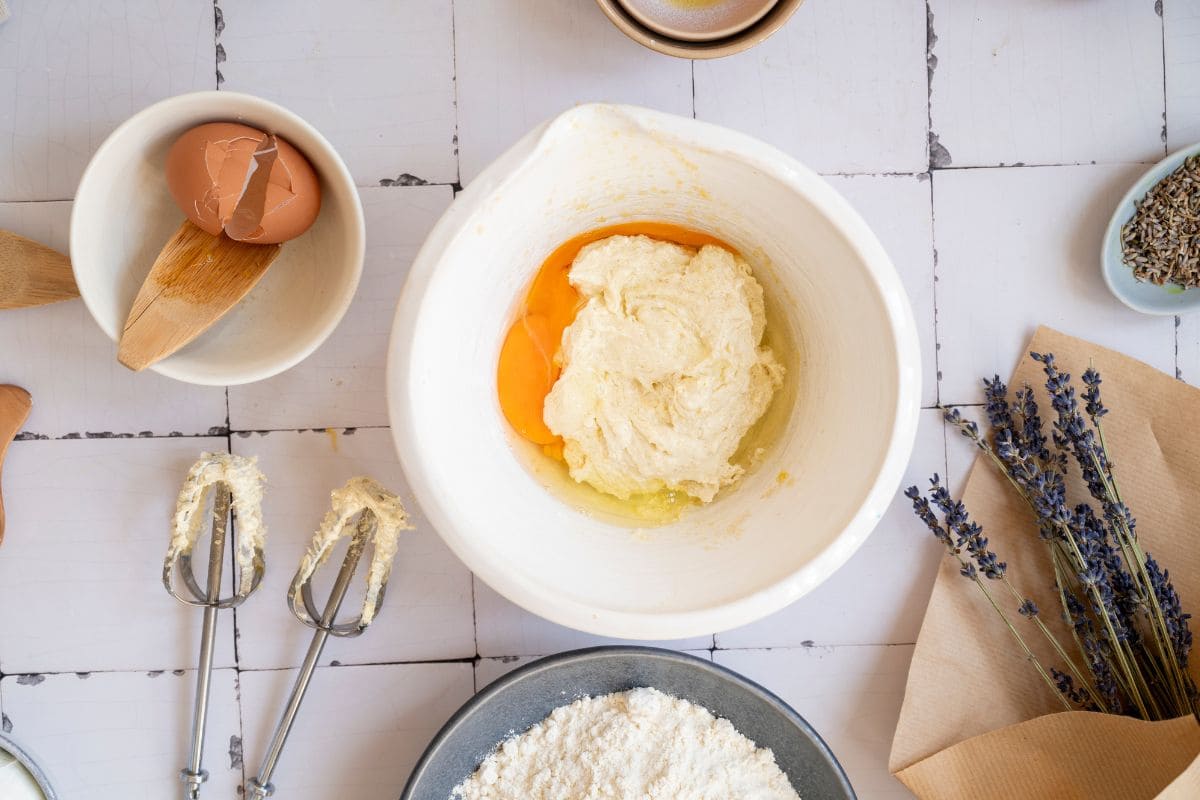 Eggshell and eggs in a bowl, baking beaters with batter, lavender flowers, and baking supplies on a white tiled surface.
