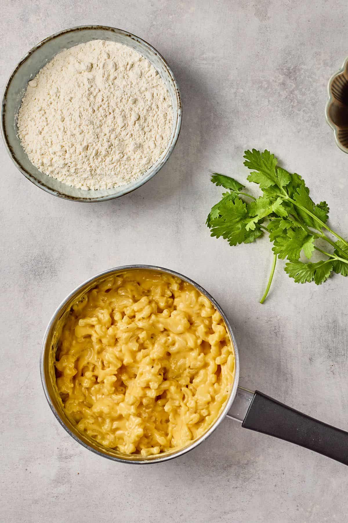 Cheddar mac and cheese in pot with fresh cilantro and baking ingredients on neutral background.