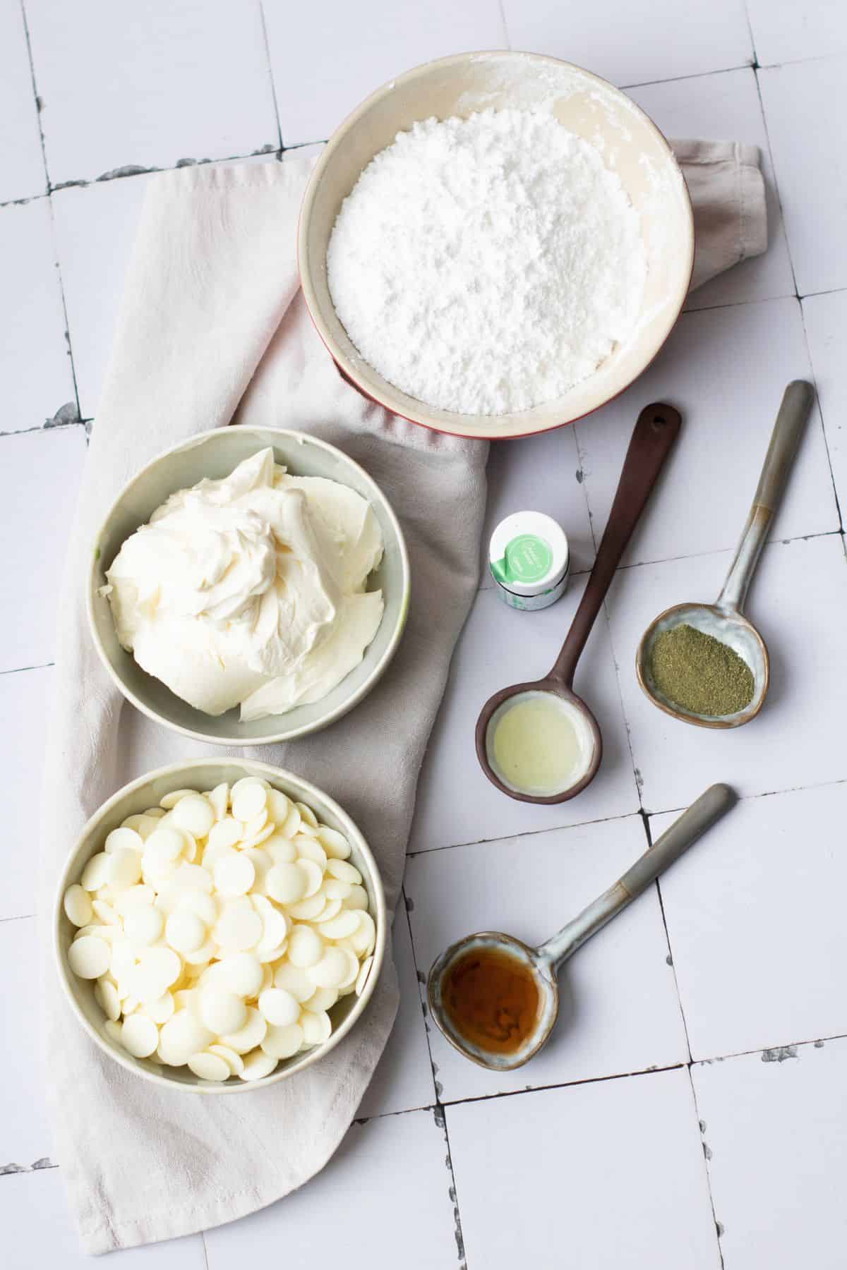 Baking ingredients including white chocolate, powdered sugar, butter, and flavorings arranged on a distressed white table.
