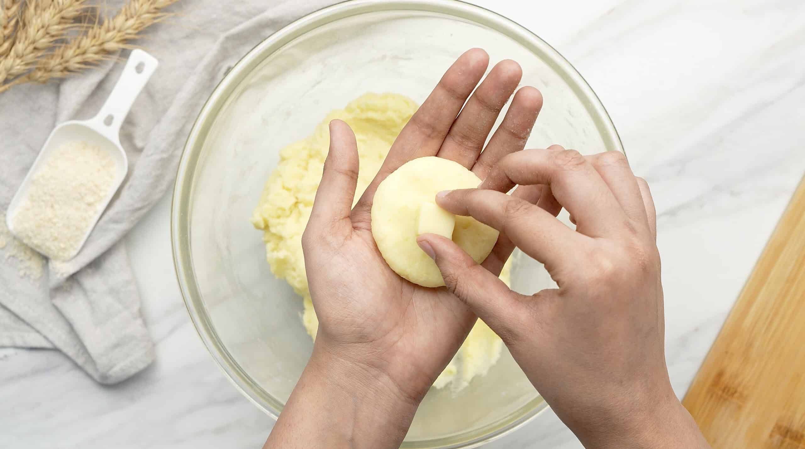 Close-up of hands shaping cookie dough in a bowl, with baking ingredients nearby.