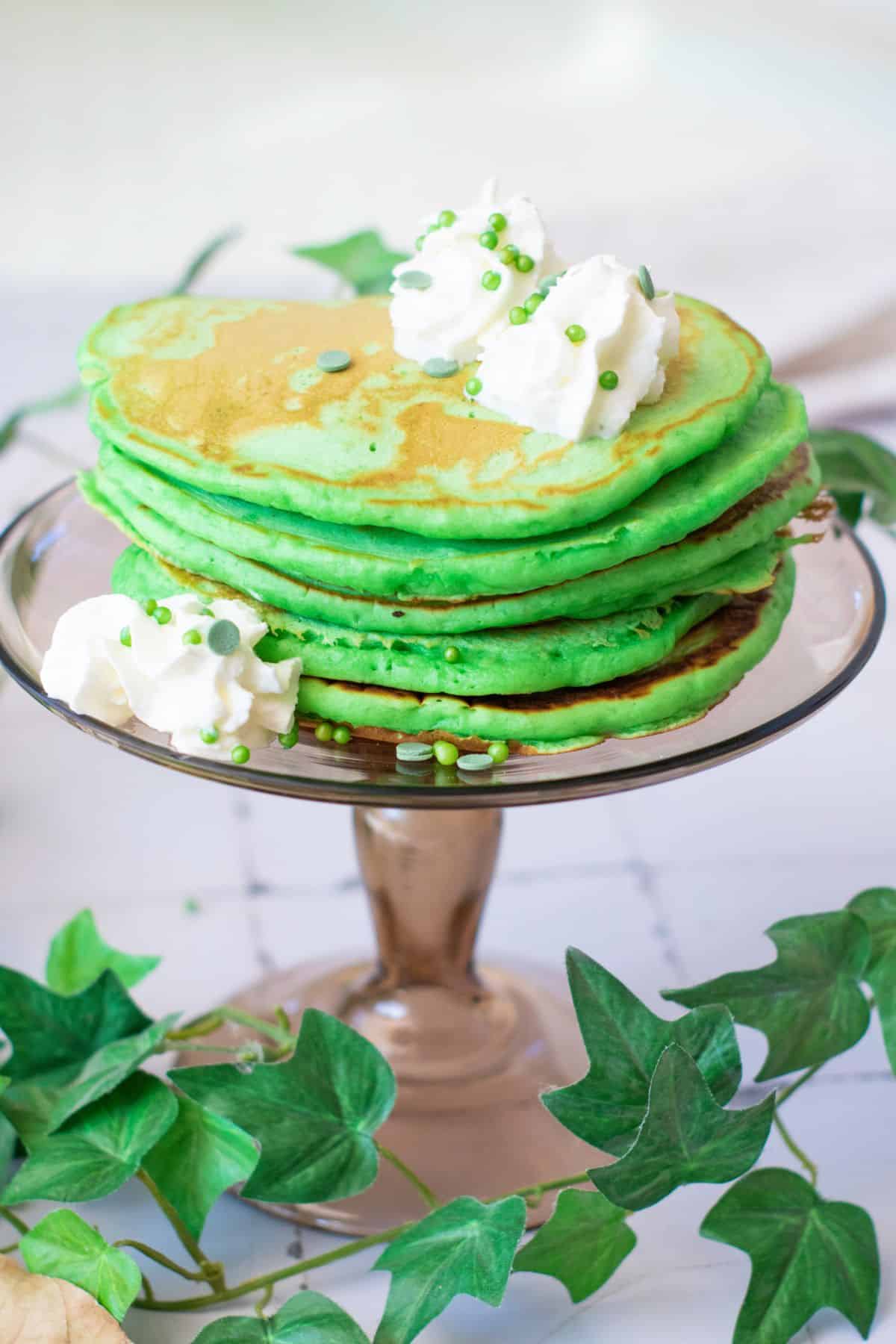 Vibrant green pancakes topped with whipped cream and decorative sprinkles, served on a glass cake stand.