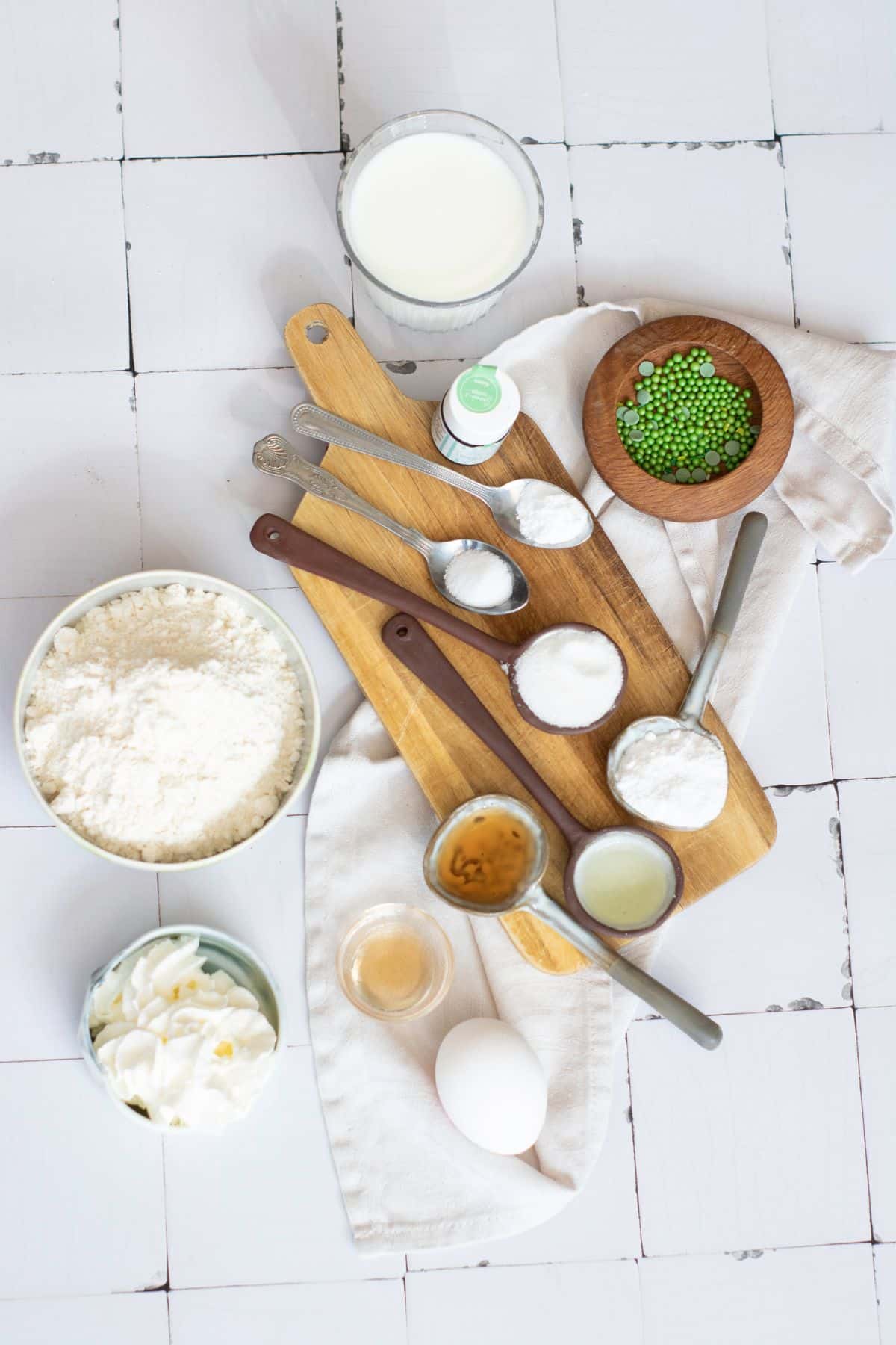 Assorted baking ingredients, measuring spoons, and a glass of milk on a rustic white kitchen table.