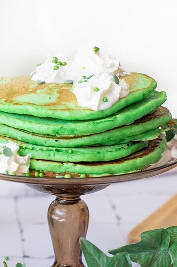 Delicious green pancakes topped with whipped cream and festive sprinkles, served on a beautiful glass cake stand.