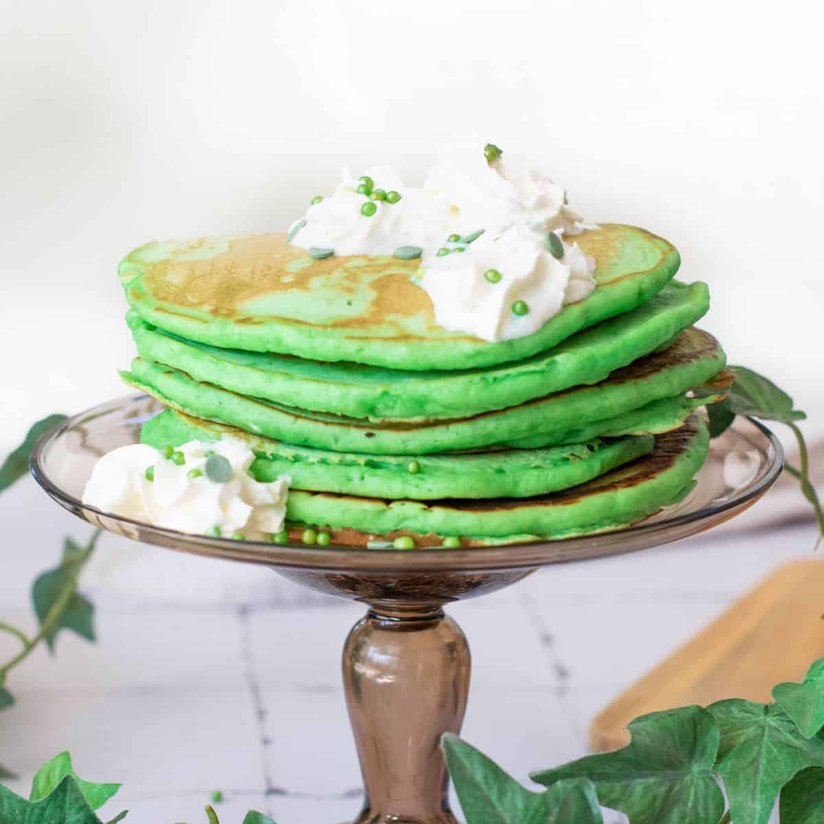 Delicious green pancakes topped with whipped cream and festive sprinkles, served on a beautiful glass cake stand.