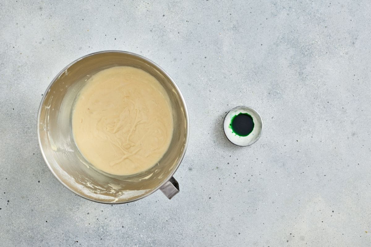 Mixing bowl with cake batter and green food coloring on a gray countertop.