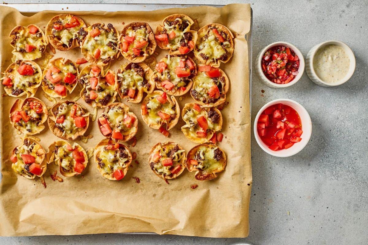 Delicious mini taco bites topped with melted cheese, diced tomatoes, served on parchment paper. Perfect for snacks or parties.
