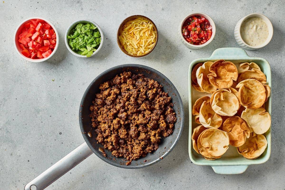 Bright, colorful taco ingredients including seasoned ground beef, tomatoes, lettuce, cheese, salsa, and sour cream arranged on a gray surface.