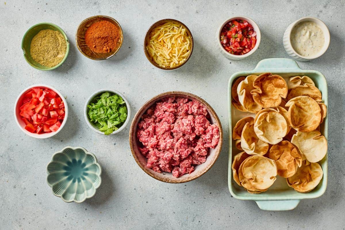 Colorful bowls with ground beef, shredded cheese, tomatoes, cilantro, and spices for tacos.